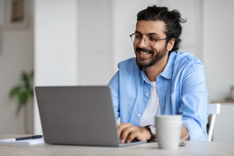 Smiling man with glasses and a beard, wearing a light blue shirt, typing on a laptop at a bright home office desk with a coffee mug beside him.
