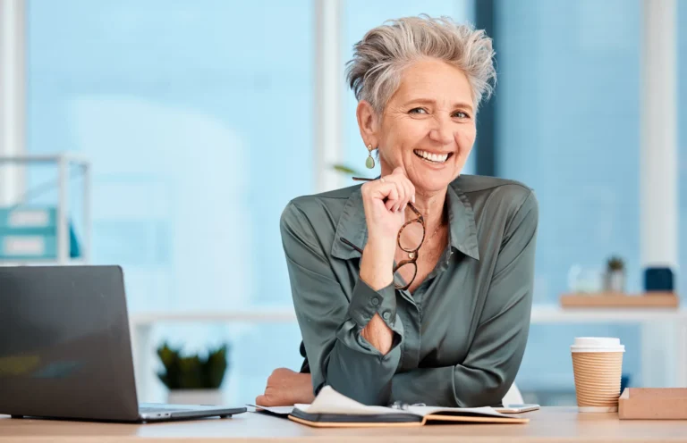 Smiling woman with short gray hair, holding her glasses, leaning on her desk in a bright office with a laptop, notebook and coffee cup.